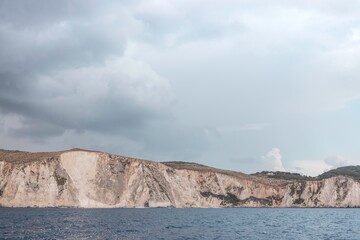 Dramatic cliffs meet the Ionian Sea under a heavy, clouded sky. The stark, white stone of the Zakynthos coastline contrasts beautifully with the deep blue of the water
