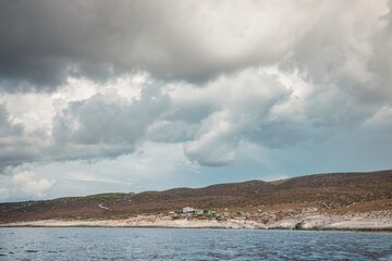 Calm blue sea meets a rocky, sparsely vegetated island in Greece. A small white house sits nestled...