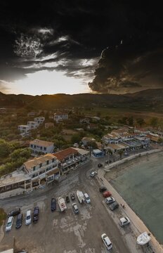 An elevated perspective captures Arkoudi Village as it meets the Ionian sea, set against a dramatic sky. Boats, cars, buildings, and distant hills fill the horizon
