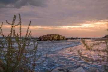 Sunlight paints the sky during dusk, casting a soft glow on the remnants of an ancient roman aqueduct. Gentle waves lap against the aged brick, creating a serene coastal view in Alanya, Turkey