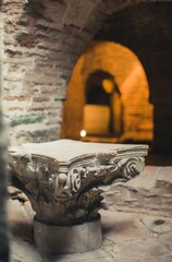 A weathered Corinthian capital stands within an ancient stone structure. A softly illuminated arched doorway provides a glimpse into further ruins, evoking history's passage