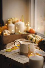 Mug of coffee standing next to the window with candles, pumpkins, book and warm blanket. Cozy home atmosphere in sunny autumn day. Cozy fall composition.