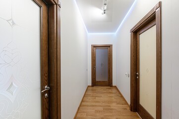 well-lit hallway with wooden doors, light walls, and a wooden floor. The ceiling has accent lighting.