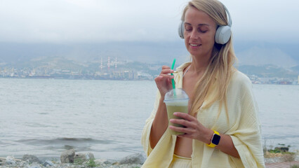 Woman drinking green smoothie with headphones by the sea