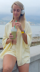Woman drinking green smoothie with headphones by the sea