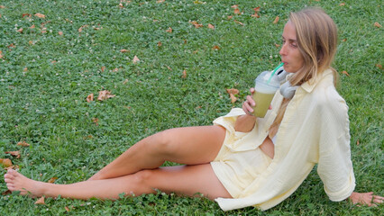 Relaxed Woman Sitting on Grass and Drinking Green Smoothie Outdoors