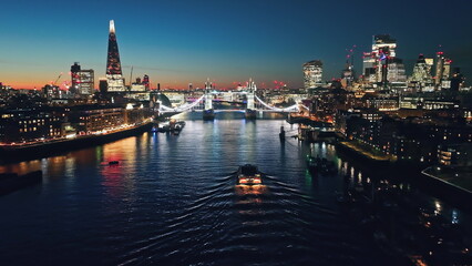 Fototapeta premium Illuminated London city skyline at sunset, Tower Bridge, The Shard, and a ferry navigating the River Thames. Night lights cityscape. United Kingdom, Europe. Travel destination. Drone flight