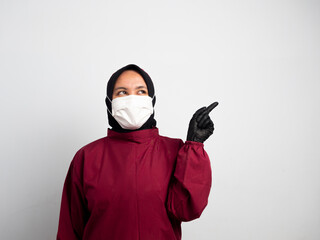 Young female nurse wearing a mask isolated on white background is surprised and pointing her finger to the side. Asian Muslim nurse wearing a red surgical gown with her finger pointing to the side.