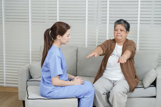 Senior asian woman lifting both arms slowly forward while sitting on sofa in rehab session guided by young female therapist wearing blue uniform observing carefully in clinic - Powered by Adobe