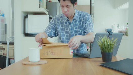 A man is walking into a kitchen with a box in his hand. The kitchen is well-equipped with a refrigerator, microwave, and oven. A laptop is on a desk in the room
