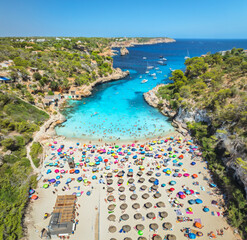 Aerial view of colorful umbrellas on crowded sandy beach, boats, hotel, rocks, swimmers in blue sea, green trees on sunny summer day. Mallorca, Balearic Islands, Spain. Tropical top drone view. Travel
