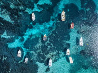 Aerial top view from drone of boats floating in shallow turquoise sea above rocky seabed with sunlight reflections, calm summer scene near Mallorca, Balearic Islands, Spain. Colorful seascape