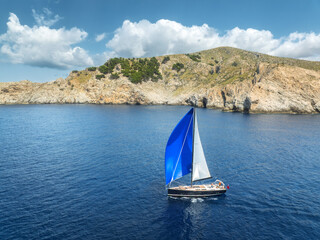 Aerial drone view of sailboat with blue and white sail cruising on deep blue Mediterranean sea near rocky coastline and cliffs. Scenic summer landscape in Mallorca, Balearic Islands, Spain. Top view