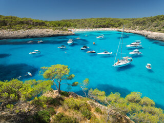 Aerial top view from drone of yachts and boats anchored in turquoise sea bay surrounded by rocky coastline and pine forest, summer vacation scene near Cala Mondrago, Mallorca, Balearic Islands, Spain