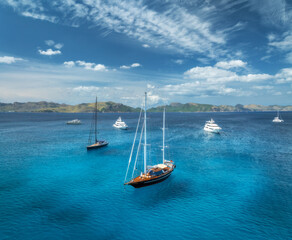 Aerial drone view of yachts and sailboats anchored in crystal clear turquoise sea near scenic coast and mountains under sky with clouds. Mallorca, Balearic Islands, Spain. Mediterranean summer travel
