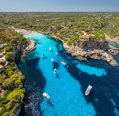 Aerial top view from drone of yachts and boats floating in narrow turquoise sea bay with rocky cliffs, green trees, sandy beach and swimmers, summer vacation scene. Mallorca, Balearic Islands, Spain