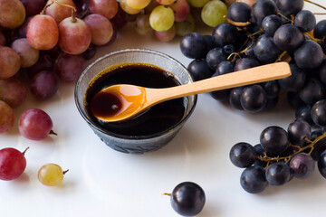 Homemade grape molasses is designed in a glass bowl with various fresh grapes on a white surface.