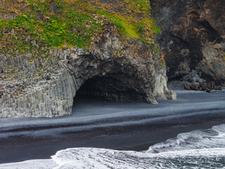 Hexagonal basalt columns and a cave at Reynisfjara beach, with green vegetation above, dark volcanic sand, and ocean waves creating a striking contrast.