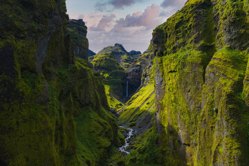 A moss covered canyon in Iceland with steep cliffs, a winding stream, and a distant waterfall under a partly cloudy sky and natural light.