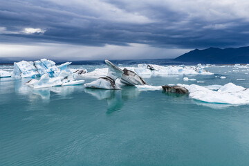 Turquoise water reflects an overcast sky as white and blue icebergs float in Jokulsarlon glacial...