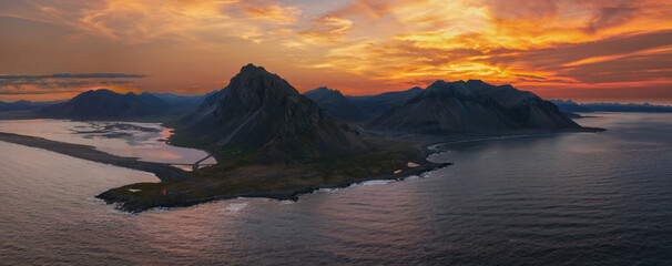 Aerial view of Iceland's Stokksnes Peninsula featuring jagged mountain peaks, a black sand beach, calm Atlantic waters, and a fiery sunset sky.