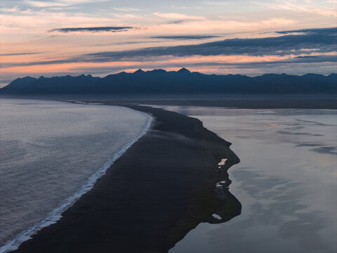 Aerial view of a black sand beach in Iceland with calm waters, jagged mountains, and a pastel sunset sky creating a dramatic landscape.