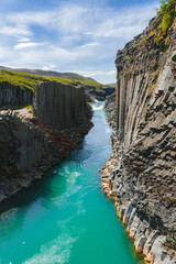 Studlagil Canyon features towering basalt columns, a vibrant turquoise river, lush green...