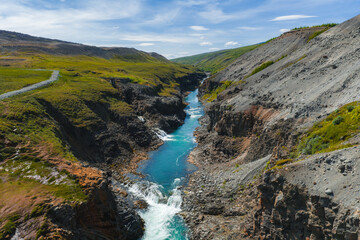 Turquoise river flows through Studlagil Canyon, flanked by basalt formations, green hills, and...
