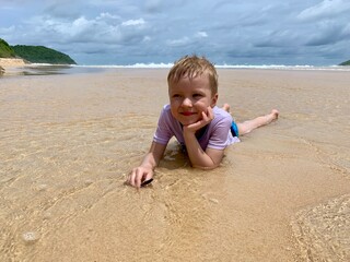 oyful little boy lying on warm tropical beach sand, smiling under the sun. Summer vacation,...
