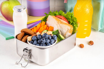 School healthy food for children. Preparing homemade balanced healthy lunch box for schooler, with sandwich, fruits, berries, juice, nuts. Ingredients and foods with lunch box on white table