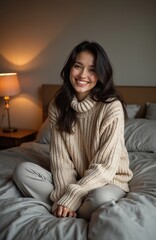 Fototapeta premium Young woman with dark hair wearing chunky knit beige sweater, light grey pants sits on bed with grey linens. Smiles warmly, with cozy loft bedroom featuring brown, grey tones in background. Soft