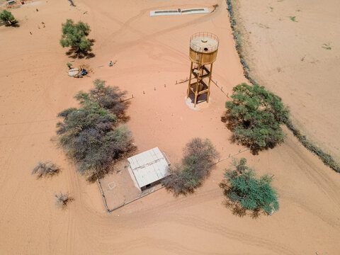 Aerial view of a water tower standing tall amidst the sandy expanse, with a building nestled among sparse trees, painting a stark contrast of civilization and nature, Khol Khol, Louga Region, Senegal.
