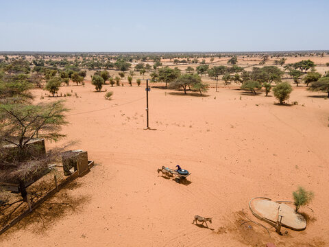 Aerial view of arid lands punctuated by sporadic trees, a simple structure, and individuals on donkey-drawn carts under a vast sky, Khol Khol, Louga Region, Senegal.