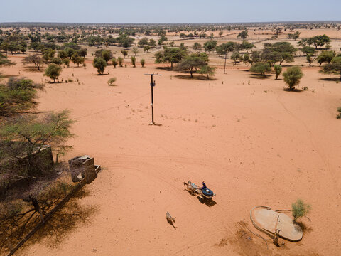 Aerial view of the vast, terracotta landscape dotted with sparse trees, a lone utility pole standing sentinel, and a small structure with a figure nearby, Khol Khol, Louga Region, Senegal.