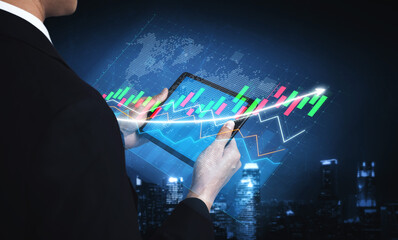A businessman stands in front of a city skyline at night, using a digital tablet to analyze...