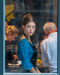 Fototapeta premium Young waitress in blue blouse seen through restaurant window, busy kitchen atmosphere and candid urban service moment
