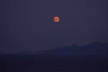In Kavala, Greece, the sun sets in warm golden tones as a soft pink moon rises, casting a gentle glow over the calm sea. The city’s silhouette fades into twilight.