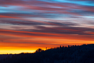 A dramatic sunrise paints the sky with vibrant red, orange, and blue hues above the silhouetted hills of the German Black Forest. The majestic clouds create a mesmerizing backdrop to the peaceful land
