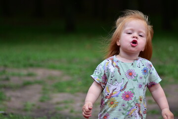 Red-haired toddler in unicorn shirt and striped leggings runs outdoors on grassy park area, hair...