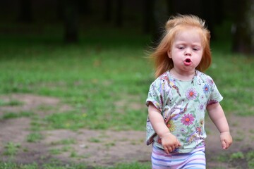 Red-haired toddler in unicorn shirt and striped leggings runs outdoors on grassy park area, hair...