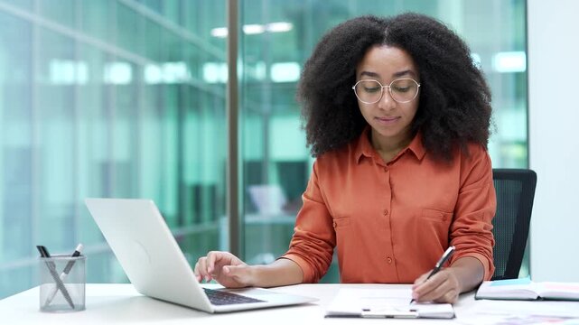 Young african american businesswoman fills out documents with a pen using laptop computer sitting at workplace in business office. female financier doing paperwork, financial report, engaged in accoun