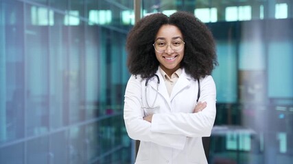 Portrait of smiling african american female doctor in white coat looking at camera in hospital clinic. Happy young black woman medical worker physician poses standing crossed arms in modern office - Powered by Adobe