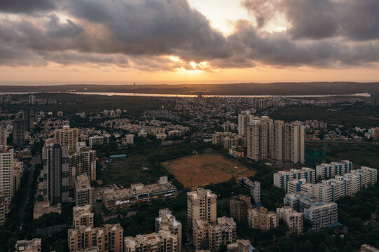 Aerial view of urban sprawl contrasting with a glimmering sunset over the distant Thane Creek, casting shadows on the city's architecture, Mumbai, Maharashtra, India.