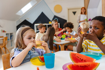 Diverse preschool children enjoying fresh watermelon at a table in a bright classroom, developing...