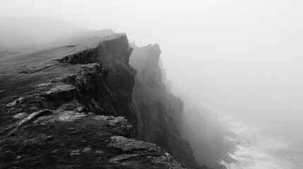 Dramatic black and white view of rugged coastal cliffs disappearing into dense fog with crashing waves below