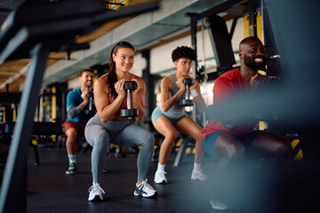 Happy athlete and her friends using hand weights while exercising squats in gym.
