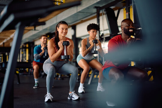 Happy athlete and her friends using hand weights while exercising squats in gym. - Powered by Adobe