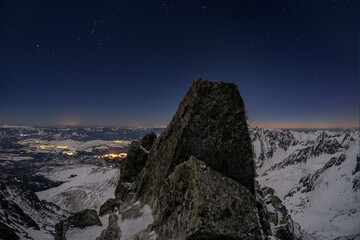 Aerial view of a craggy peak under a starlit sky, with the distant town lights twinkling like scattered jewels, Koncista, PreÅ¡ov Region, Slovakia.