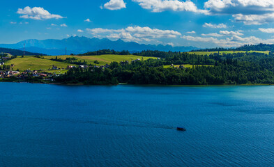 Wide view of Czorsztyn Lake with a motorboat crossing the water. Tatra Mountains and green hills stretch across the horizon.