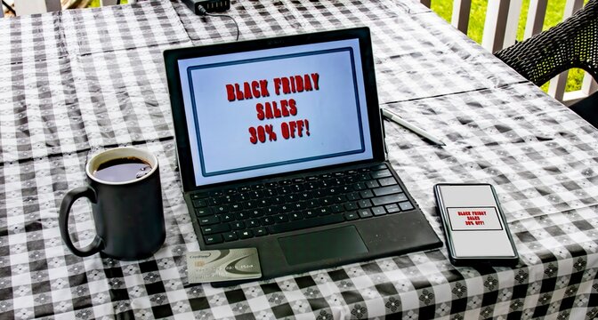 A black checkered table is set up outdoors featuring a laptop displaying a Black Friday sale announcement, a coffee cup, and a smartphone nearby. The scene suggests a cozy shopping experience.